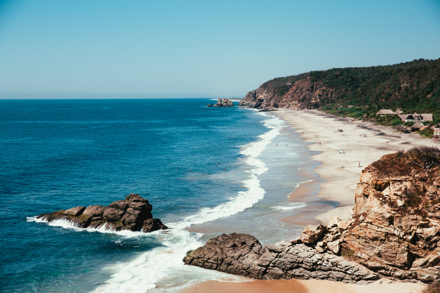 Scenic photo of coastline with bright blue ocean on the left and the shore meeting cliffs on the right. There is one large rock at the edge of the shoreline on the bottom left of the photo. 